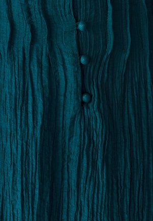 Tealowa, teksturowana tkanina z pionowymi fałdami; zawiera cztery okrągłe, teksturowane guziki wzdłuż środka. Miękki, pomarszczony wygląd.