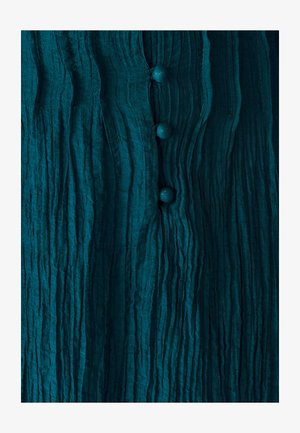 Tealowa, teksturowana tkanina z pionowymi fałdami; zawiera cztery okrągłe, teksturowane guziki wzdłuż środka. Miękki, pomarszczony wygląd.