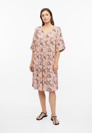 Femme debout dans une robe rose à motifs longueur genou avec des manches larges et des sandales noires à bouts ouverts sur fond blanc.