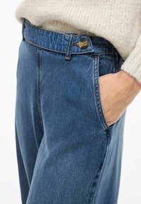 Visoke plave traperice s ravnim krojem, koje imaju mesing dugme, prednje džepove i svijetli pranje.