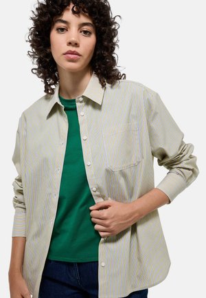 Chemise à rayures dans des tons de jaune clair, bleu et blanc, avec une poche poitrine. Portée par-dessus un t-shirt vert, avec des manches longues et une coupe décontractée.