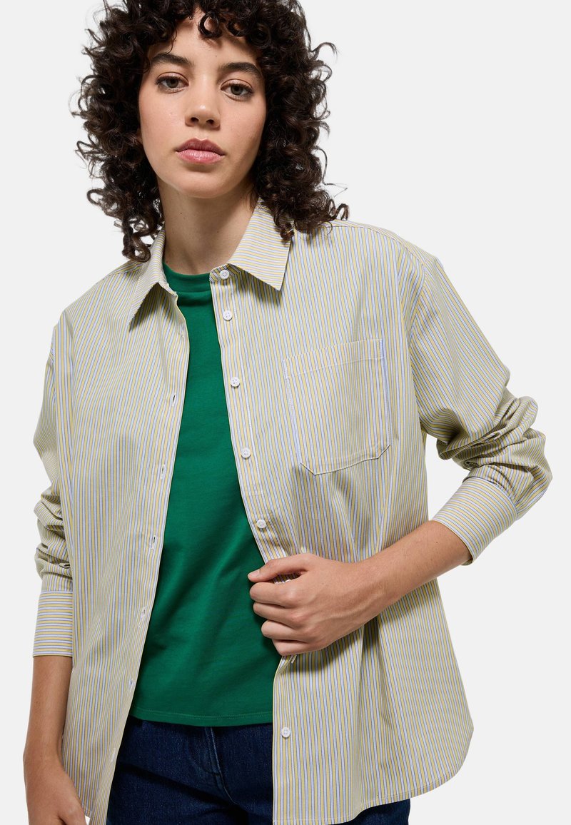 Chemise à rayures dans des tons de jaune clair, bleu et blanc, avec une poche poitrine. Portée par-dessus un t-shirt vert, avec des manches longues et une coupe décontractée.