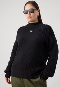 Pull en maille côtelée noir avec un col montant, présentant une patch logo. Le mannequin porte des lunettes de soleil sombres. Tissu texturé avec des manches larges.