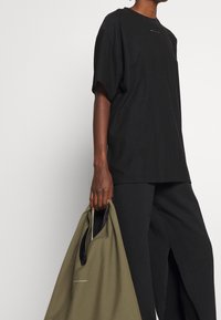 Svart oversized t-shirt med rund halsringning och korta ärmar, kombinerad med svarta vida byxor. Modellen håller en olivgrön tote-väska.