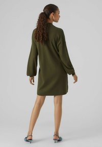 Vestito a maglia verde oliva con maniche lunghe e a sbuffo, dal taglio rilassato, con un collo alto e spacchi laterali. Indossato con tacchi blu.