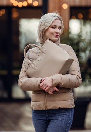 Beige Handtasche mit abgerundeten Griffen, glatter Oberfläche und eingeprägtem Logo. Kombiniert mit einer hellbraunen Steppjacke und blauer Jeans.