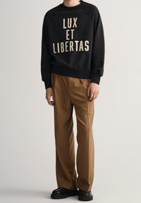Felpa nera con la scritta "LUX ET LIBERTAS" in lettere crema, abbinata a pantaloni beige plissettati e scarpe nere con suola spessa.