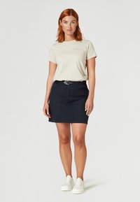 Beige tričko s logem, doplněné tmavě modrou džínovou sukní s páskem a kapsami, nošeno s bílými teniskami. Jednoduchý a neformální design.