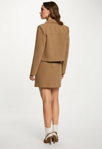 Vrouw die zijwaarts staat in een beige cropped jack en bijpassende rok, met witte sokken en bruine hoge hakken tegen een effen achtergrond.