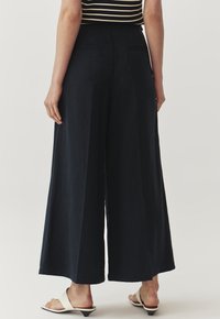 Marineblauwe brede broeken met een gladde textuur, featuring een hoge taille en twee achterzakken, gecombineerd met eenvoudige sandalen.