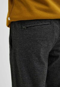 Černé a šedé kostkované kalhoty s texturovanou látkou, s kapsou na zip na zadní straně a nad nimi hořčicově žlutá košile.