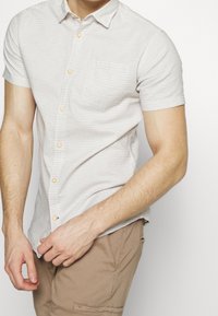 Homme portant une chemise blanche à manches courtes avec des rayures subtiles et un pantalon beige, ajustant l'ourlet de la chemise avec les deux mains.