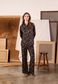 Navy and gold floral patterned shirt and pants set, made from soft fabric, featuring a collared neckline and relaxed fit. Model stands against art backdrop.