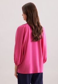 Blusa rosa in seta con una vestibilità ampia, dettaglio delle spalle arricciato e lunghe maniche a sbuffo. Il tessuto ha una texture liscia.