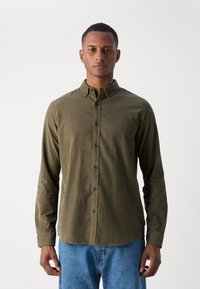 Olivenfarget langermet skjorte med knappekrage, frontknapper og avslappet passform. Kombinert med blå denimjeans.
