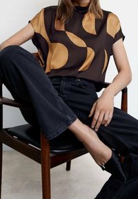 Personne assise sur une chaise en bois, portant un pantalon noir, des chaussures noires à bout pointu, et un chemisier à manches courtes avec de grandes formes abstraites beiges.