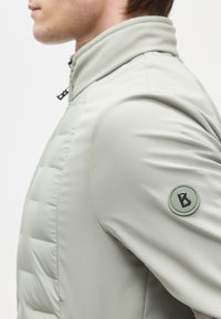 Vue latérale en gros plan d'une personne portant une veste matelassée gris clair avec un col montant et un patch logo rond sur la partie supérieure de la manche.