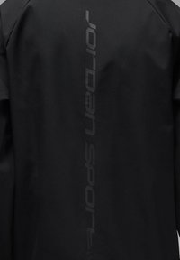 Chaqueta negra con una textura suave y un logo vertical "Jordan Sport" en un tono más oscuro en la parte posterior. Diseño minimalista y sin detalles adicionales.