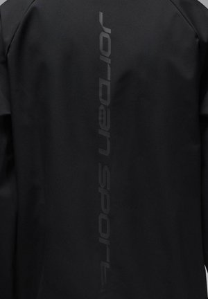 Giacca nera con una texture liscia e un logo verticale "Jordan Sport" di tonalità più scura sul retro. Design minimalista e senza dettagli aggiuntivi.