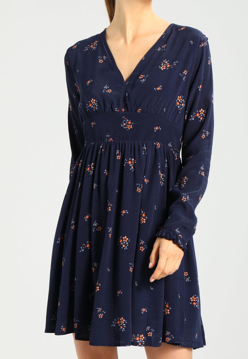 Femme portant une robe bleu marine à manches longues avec de petits motifs floraux orange et blanc et une taille froncée.