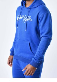 Uomo che indossa un felpa azzurra brillante e pantaloni tuta abbinati con testo corsivo bianco sul petto, in piedi contro uno sfondo bianco semplice.