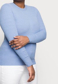 Hellblauer Strickpullover mit Rippstruktur und langen Ärmeln, mit Rundhalsausschnitt. Getragen zu weißen Hosen.