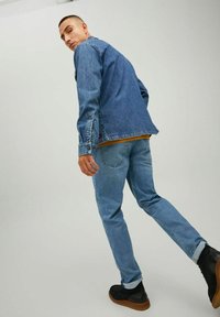 Chaqueta de mezclilla en azul desgastado, con aberturas laterales. La modelo lleva una camisa mostaza debajo, combinada con jeans de un azul claro y zapatos negros.