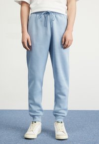 Světle modré tepláky s elastickým pasem a šňůrkou, mají stažené kotníky a hladkou, jemnou texturu. Kombinované s bílými teniskami.