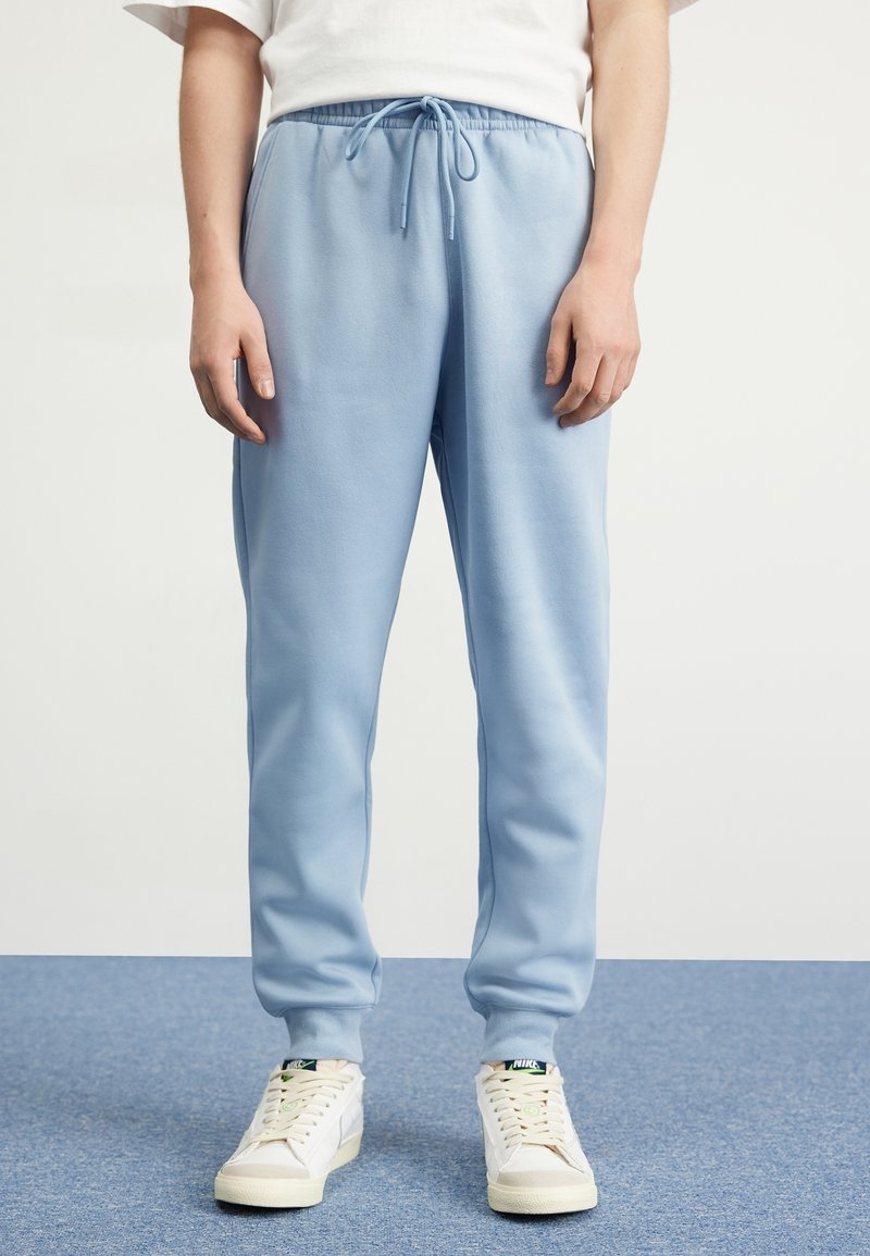 Světle modré tepláky s elastickým pasem a šňůrkou, mají stažené kotníky a hladkou, jemnou texturu. Kombinované s bílými teniskami.