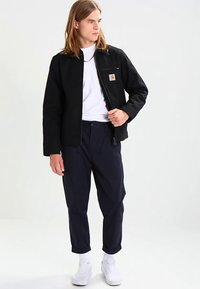 Giacca nera con zip frontale, toppa con logo, abbinata a una t-shirt bianca e pantaloni blu navy. Indossata con scarpe da ginnastica bianche.