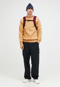 Felpa beige con polsini a costine, pantaloni cargo scuri con tasche con zip, zaino bordeaux e berretto blu navy con testo bianco. Scarpe bianche.