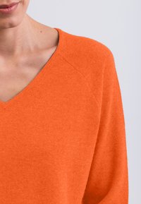 Maglione a V con maniche lunghe di un brillante arancione, realizzato in un tessuto morbido e strutturato. Presenta maniche raglan e una vestibilità rilassata.
