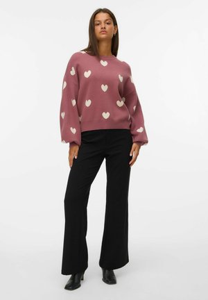 Suéter en rosa polvo con patrones de corazones crema; corte relajado, mangas largas. Pantalones acampanados negros con una textura suave y un corte a medida.