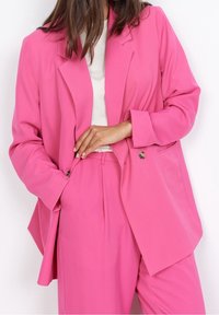 Jasnoróżowa marynarka dopasowana o luźnym kroju, z klapami i guzikami. Nosi się ją na białej bluzce, zestawiona z pasującymi różowymi spodniami.