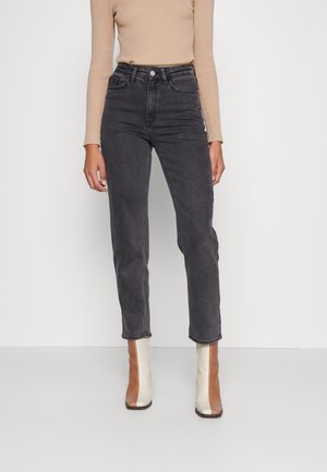 Femme portant un jean noir taille haute, un haut beige à manches longues côtelé, et des bottines bicolores marron et blanches, debout devant un fond uni.