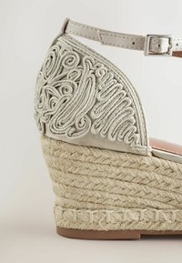 Sandale espadrille compensée avec semelle en jute tressé naturel, talon beige brodé, et bride de cheville réglable avec boucle en métal.