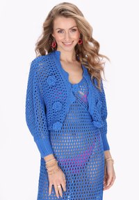 Niebieski szydełkowany kardigan z kwiatowymi akcentami, z ażurowym wzorem i długimi rękawami, na różowej bluzce. Gładka faktura z prążkowanymi mankietami.