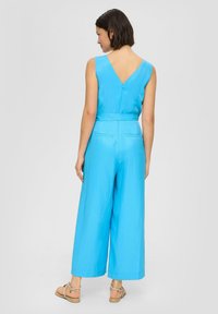 Blauwe katoenen jumpsuit met een diep V-rug, wijde pijpen, gecinchte taille en zakken. Gladde textuur en eenvoudige, strakke lijnen.