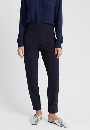 Woman wearing navy long-sleeve blouse, fitted navy ankle-length pants with cuffs, and silver snakeskin-patterned slingback flats.