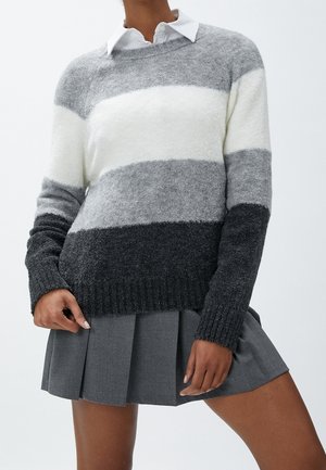 Femme portant un pull duveteux rayé gris, blanc et noir par-dessus une chemise blanche à col et une jupe plissée grise.