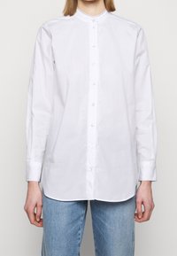 Chemise blanche à manches longues, en coton, dotée d'un col montant et d'une fermeture à boutons avec des coutures décoratives le long de la patte de boutonnage.