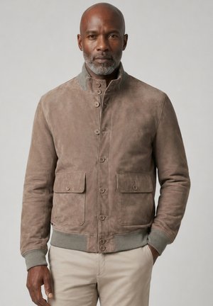 Mann trägt eine braune Wildlederjacke mit Knopfverschluss und beige Hose, steht mit einer Hand in der Tasche vor einem schlichten Hintergrund.