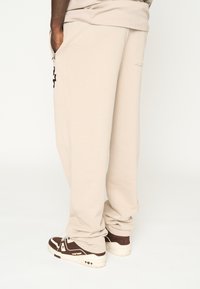 Pantalones de chándal beige con un ajuste relajado, que cuentan con bolsillos laterales y un detalle de logo negro. Combinados con zapatillas marrones y blancas.