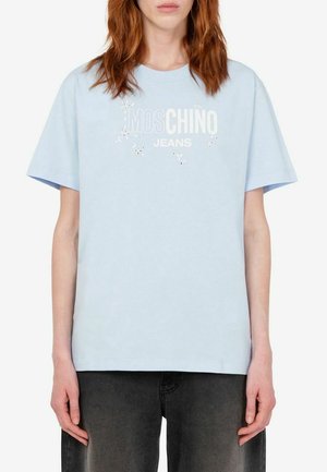 Donna con capelli rossi che indossa una t-shirt Moschino Jeans azzurro chiaro e jeans grigio scuro, in piedi davanti a uno sfondo bianco uniforme.
