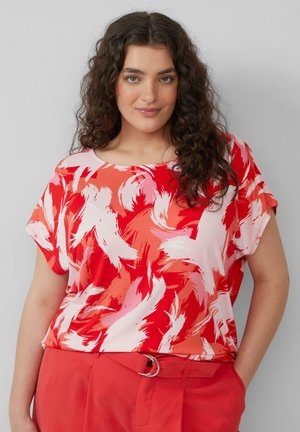 Vrouw met krullend donker haar, gekleed in een top met een abstracte print in rood en roze en rode broek met een riem, staand met één hand in de zak.