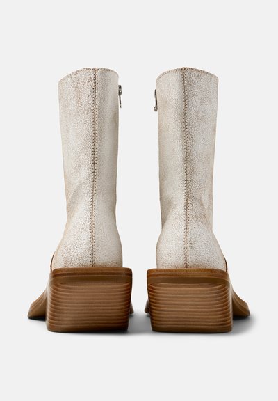 Par de botas altas de cuero texturizado color blanco roto con tacones de bloque de madera apilados, vistas desde atrás sobre un fondo blanco.