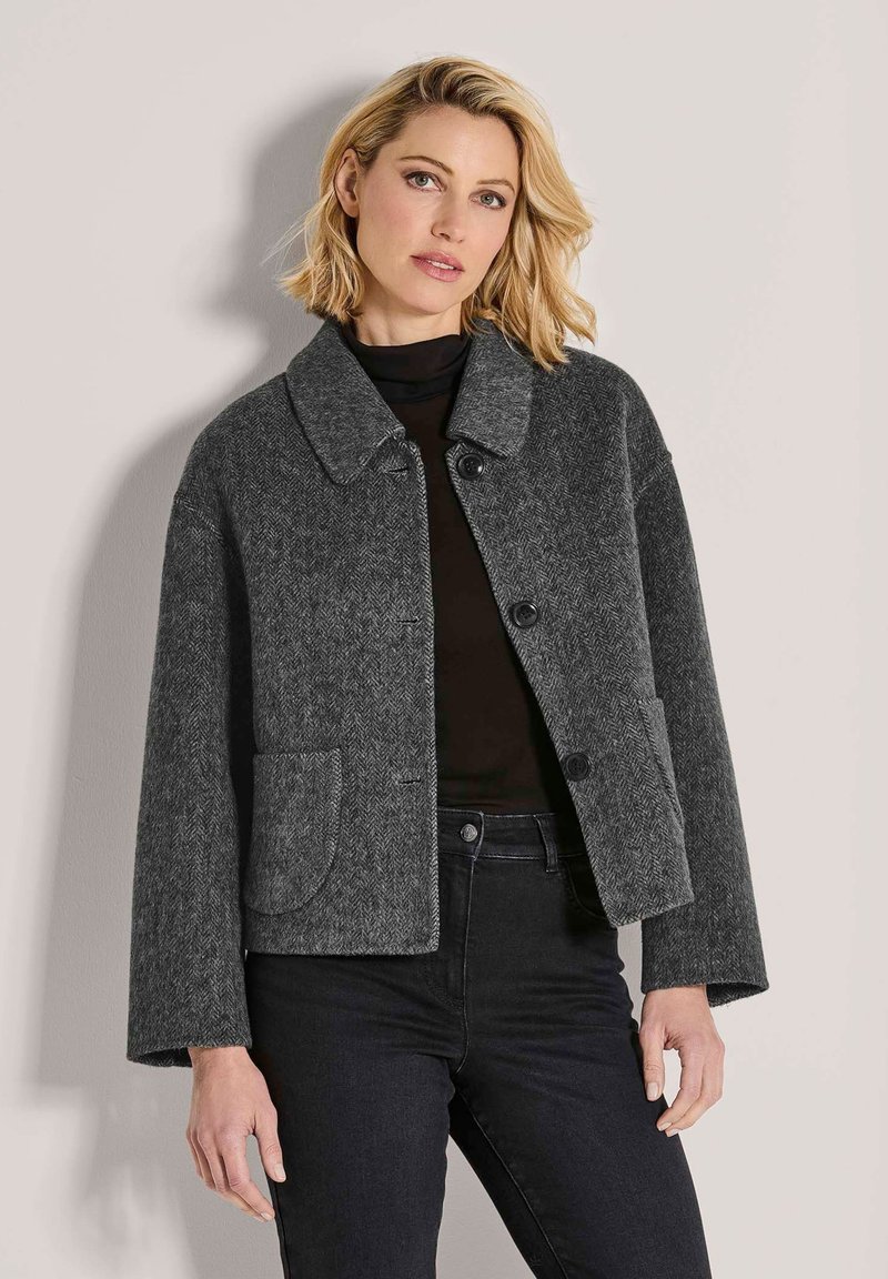 Blonde vrouw in een donkergrijze herringbone knoopjack met grote kraag, een zwarte coltrui en zwarte broek, staand tegen een effen muur.