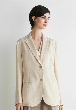 Frau mit kurzen dunklen Haaren, die ein cremefarbenes, gestreiftes Blazer mit zwei Knöpfen trägt, steht vor einer schlichten hellen Wand und blickt nach rechts.