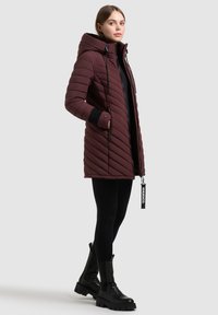 Bordo puffer jakna s chevron uzorkom, kapuljačom, crnim manžetama i zatvaranjem na patentni zatvarač, u kombinaciji s crnim gležnjačama.
