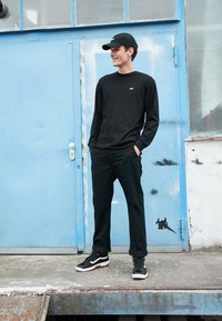 Camicia nera a manica lunga con piccolo logo, pantaloni neri e sneakers nere. In piedi di fronte a una porta blu, look streetwear casual.